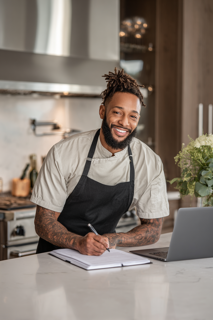 A 30-something private chef in a home kitchen with a laptop and notepad